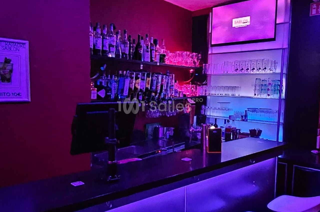 Bar avec comptoir éclairé, étagères remplies de bouteilles et verres, ambiance lumineuse violette et bleue.