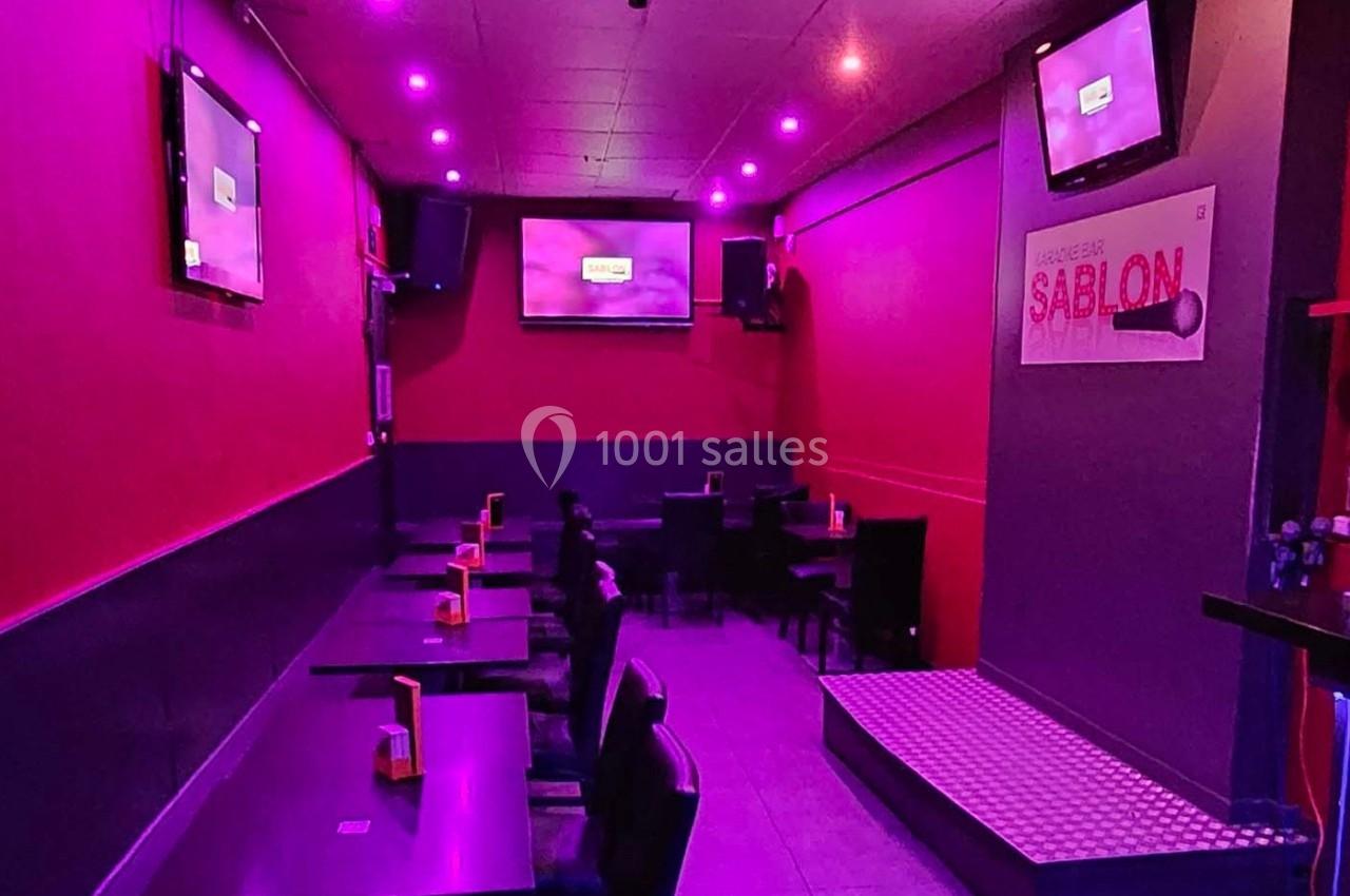 Salle de karaoké avec éclairage violet, tables alignées et écrans accrochés aux murs.