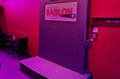 Scène de karaoké dans un bar avec éclairage rose et rouge, micro illustré sur un panneau mural.