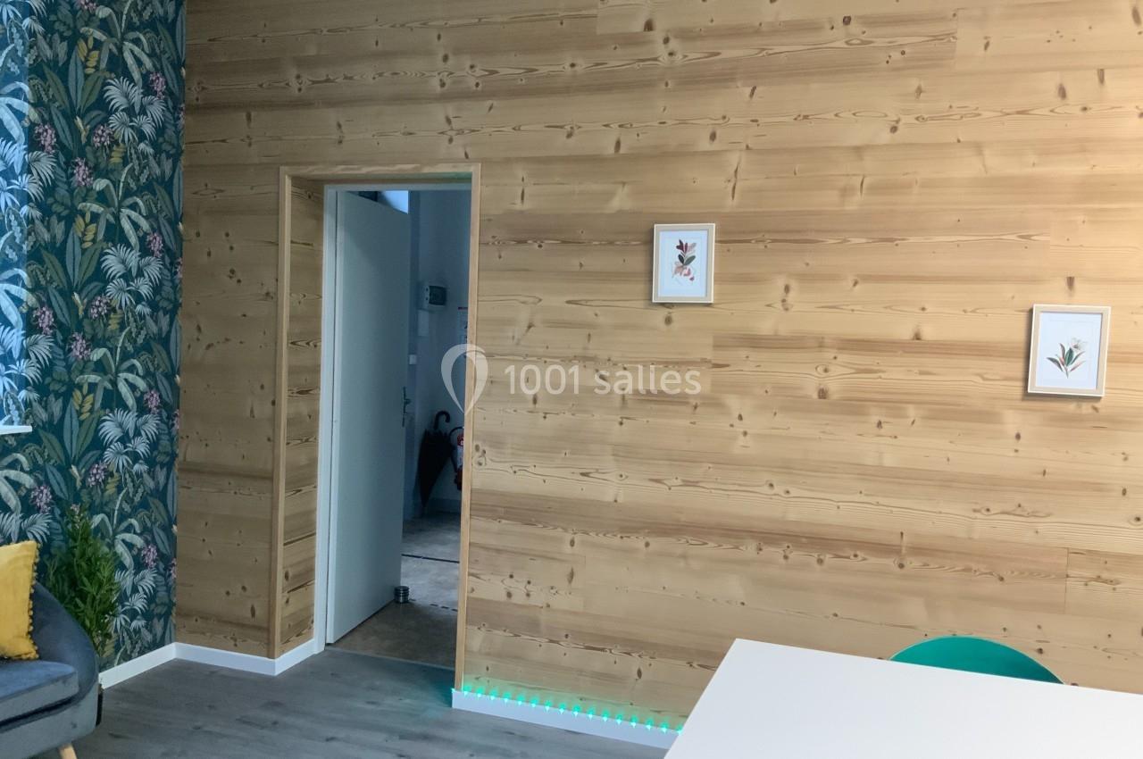 Mur en bois clair avec deux cadres décoratifs, porte ouverte sur un couloir, sol en parquet et coin de mobilier visible.
