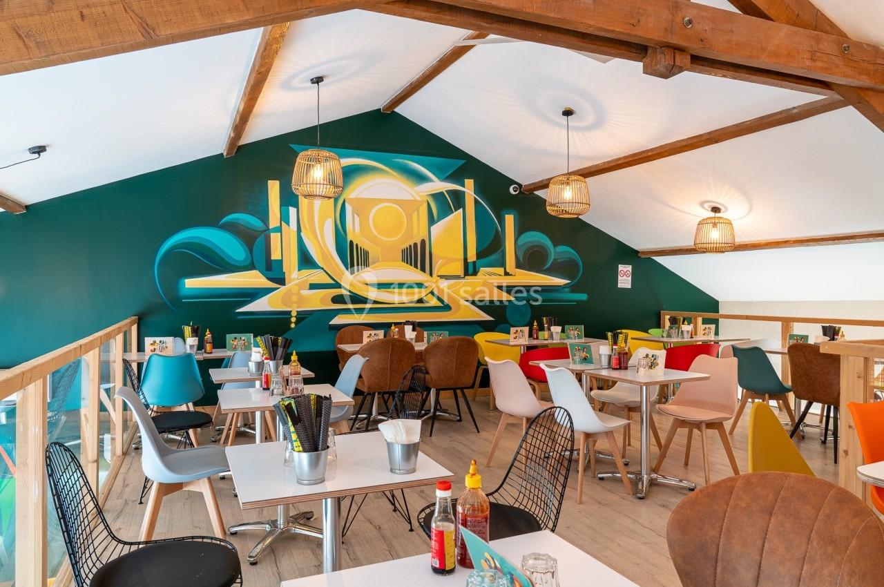 Salle de restaurant colorée avec des tables modernes, des chaises variées et une fresque murale géométrique sur fond vert.