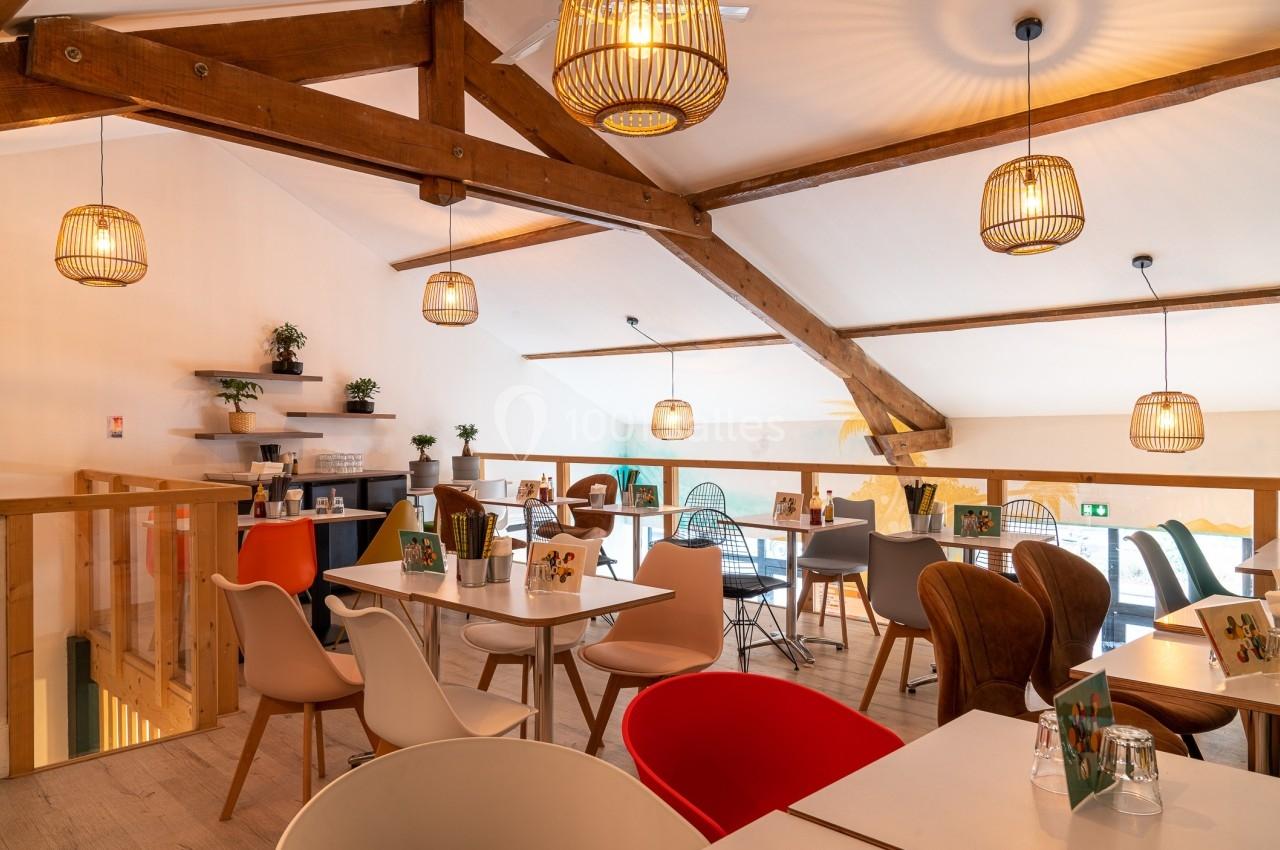 Salle lumineuse avec poutres apparentes, tables et chaises modernes, décorée de plantes et éclairée par des suspensions.