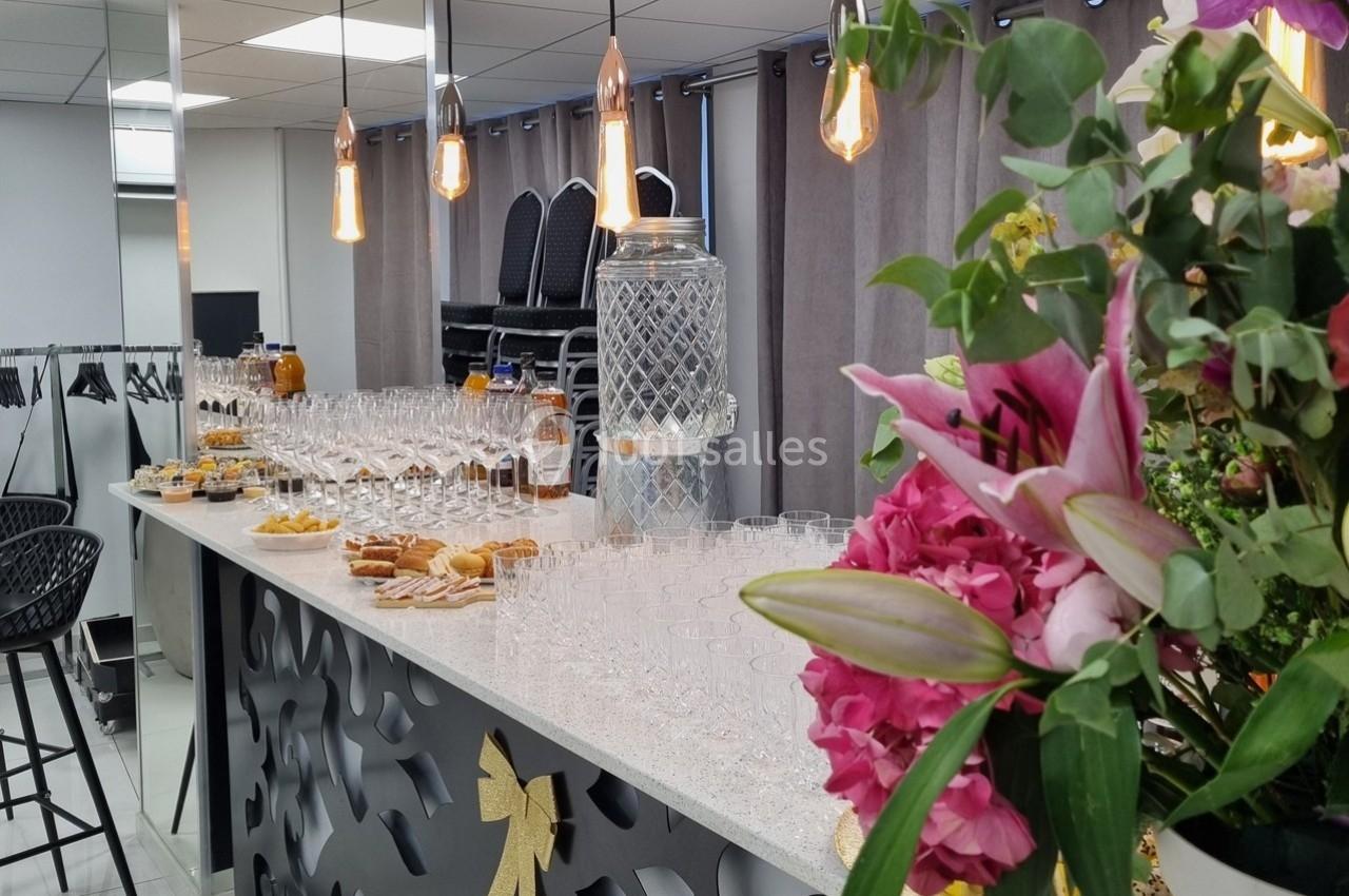 Table de réception avec verres, carafes, fleurs et collations, dans une salle lumineuse avec rideaux et chaises empilées.