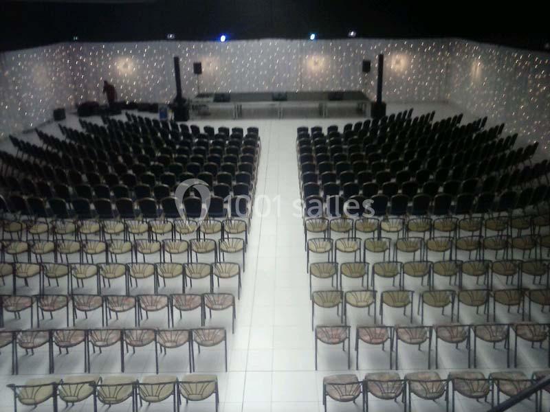 Salle de conférence avec des rangées de chaises alignées face à une scène équipée de tables et micros.