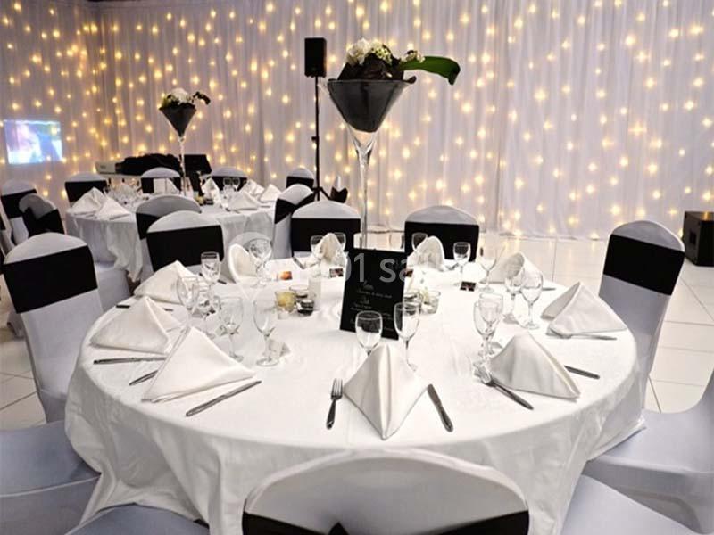 Salle de réception décorée avec des tables rondes dressées, nappes blanches et fond lumineux avec guirlandes.