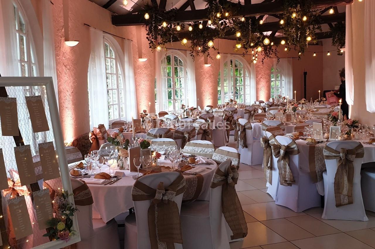 Salle de réception décorée avec des tables dressées, des chaises ornées de rubans et un plan de table à l'entrée.