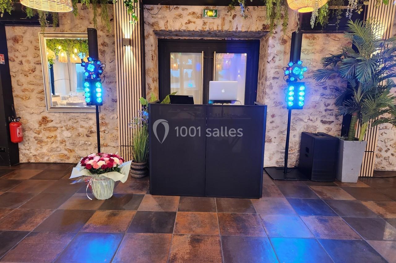 Poste de DJ avec ordinateur portable, éclairages colorés, bouquet de fleurs et décor végétal dans une salle en pierre.