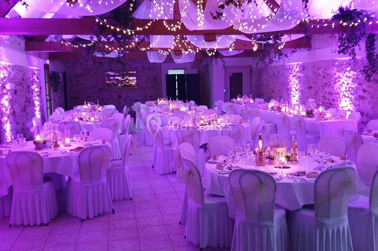 Salle de réception décorée avec des tables rondes, nappes blanches et éclairage violet, ornée de guirlandes lumineuses.