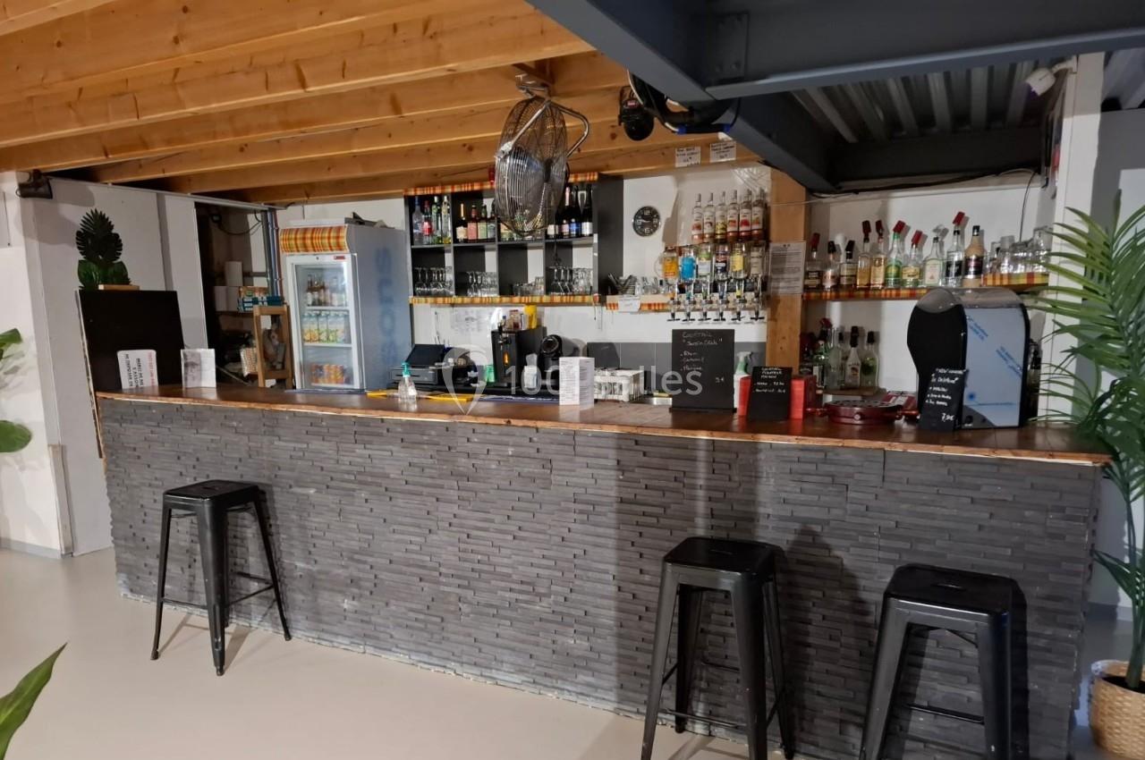 Comptoir de bar en pierre avec tabourets noirs, étagères remplies de bouteilles et réfrigérateur en arrière-plan.