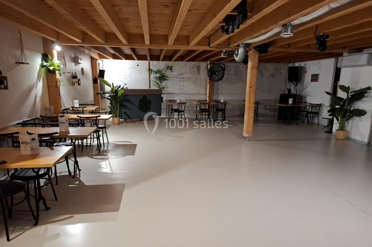 Salle lumineuse avec tables et chaises en bois, plafond en poutres apparentes, plantes et décor mural minimaliste.