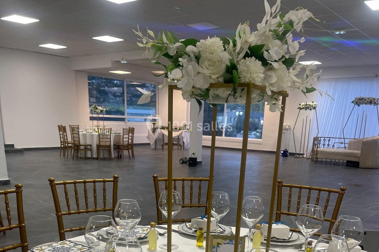 Centre de table floral blanc sur une table dressée dans une salle de réception spacieuse avec vue sur l'extérieur.