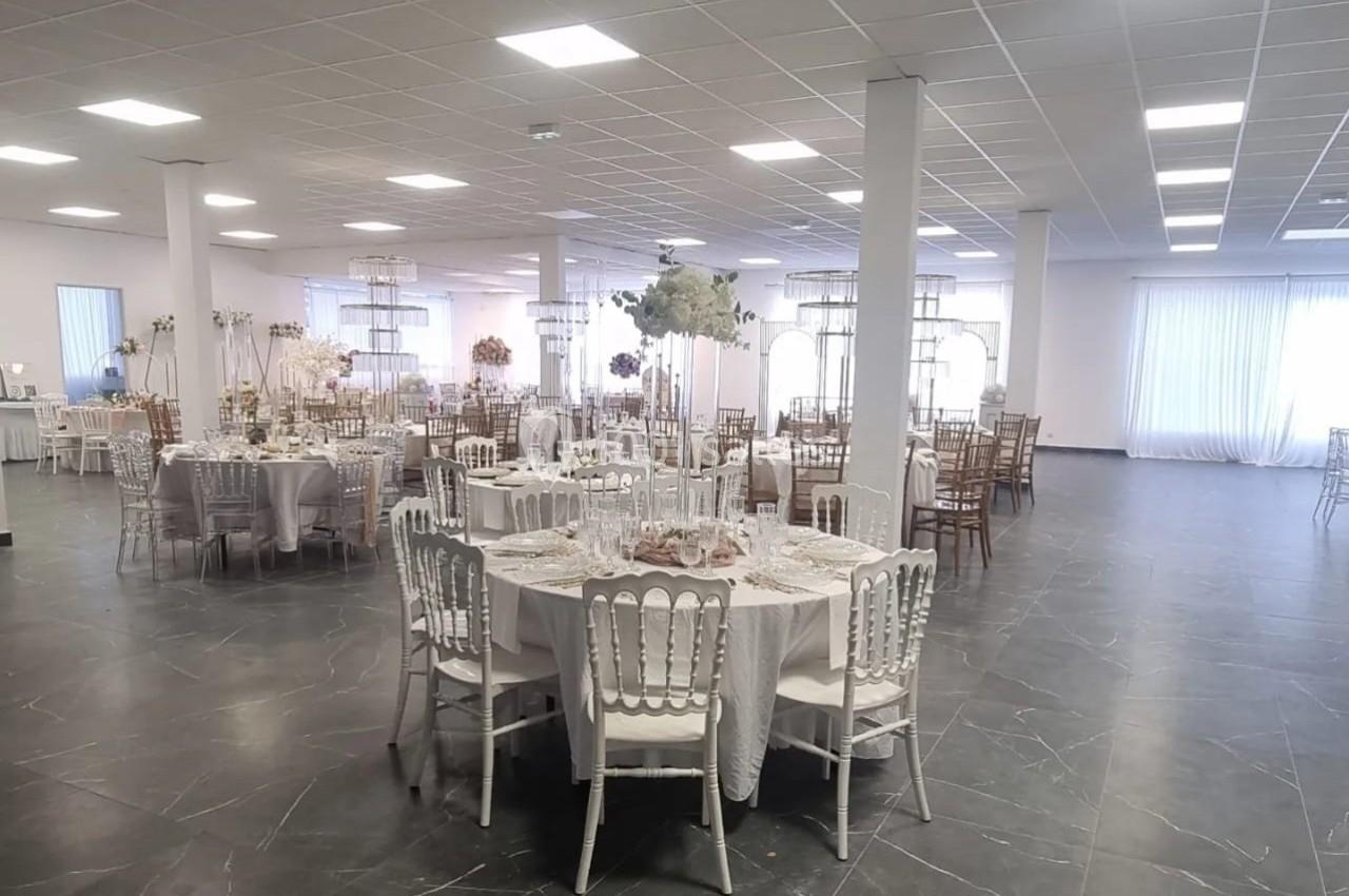Salle de réception lumineuse avec tables rondes dressées, chaises blanches et décorations florales élégantes.