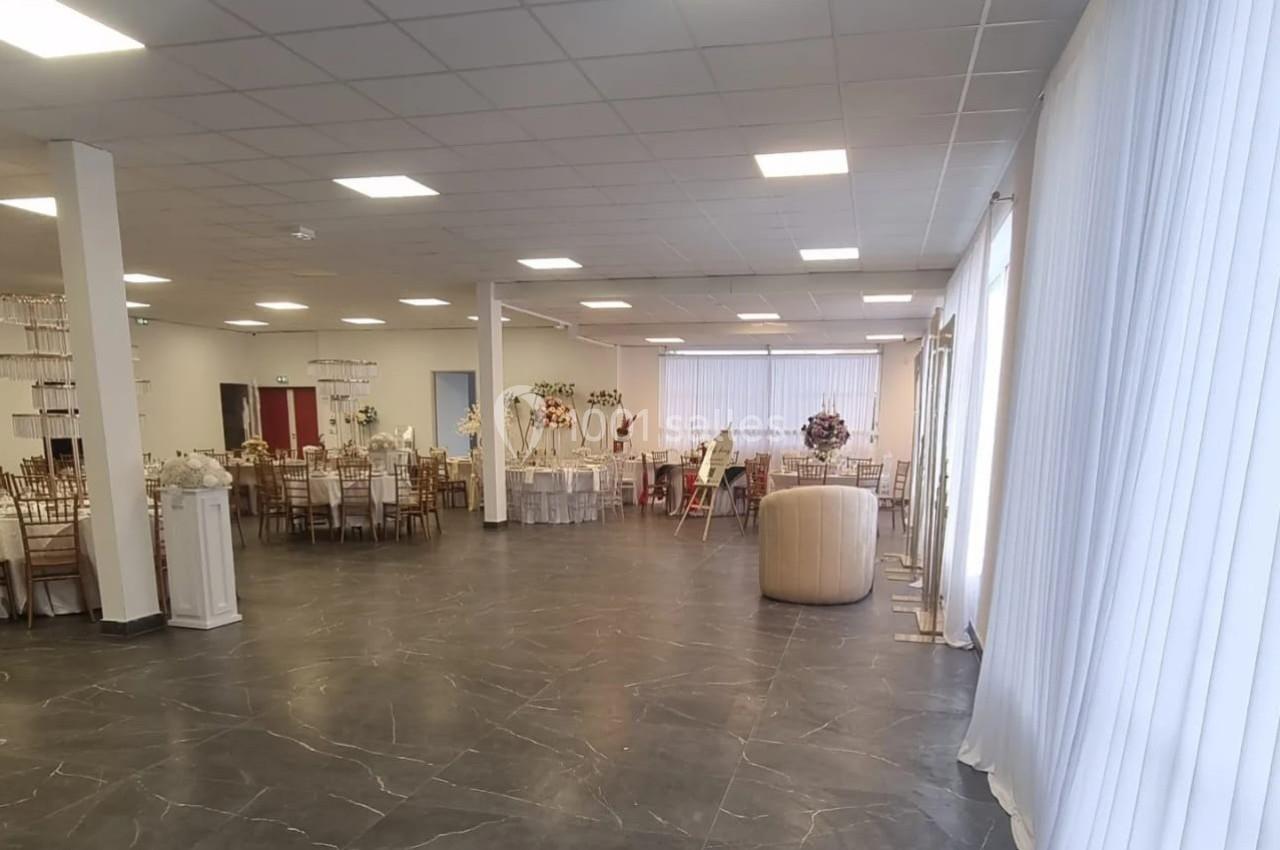 Salle de réception lumineuse avec tables décorées, chaises en bois et rideaux blancs couvrant les fenêtres.