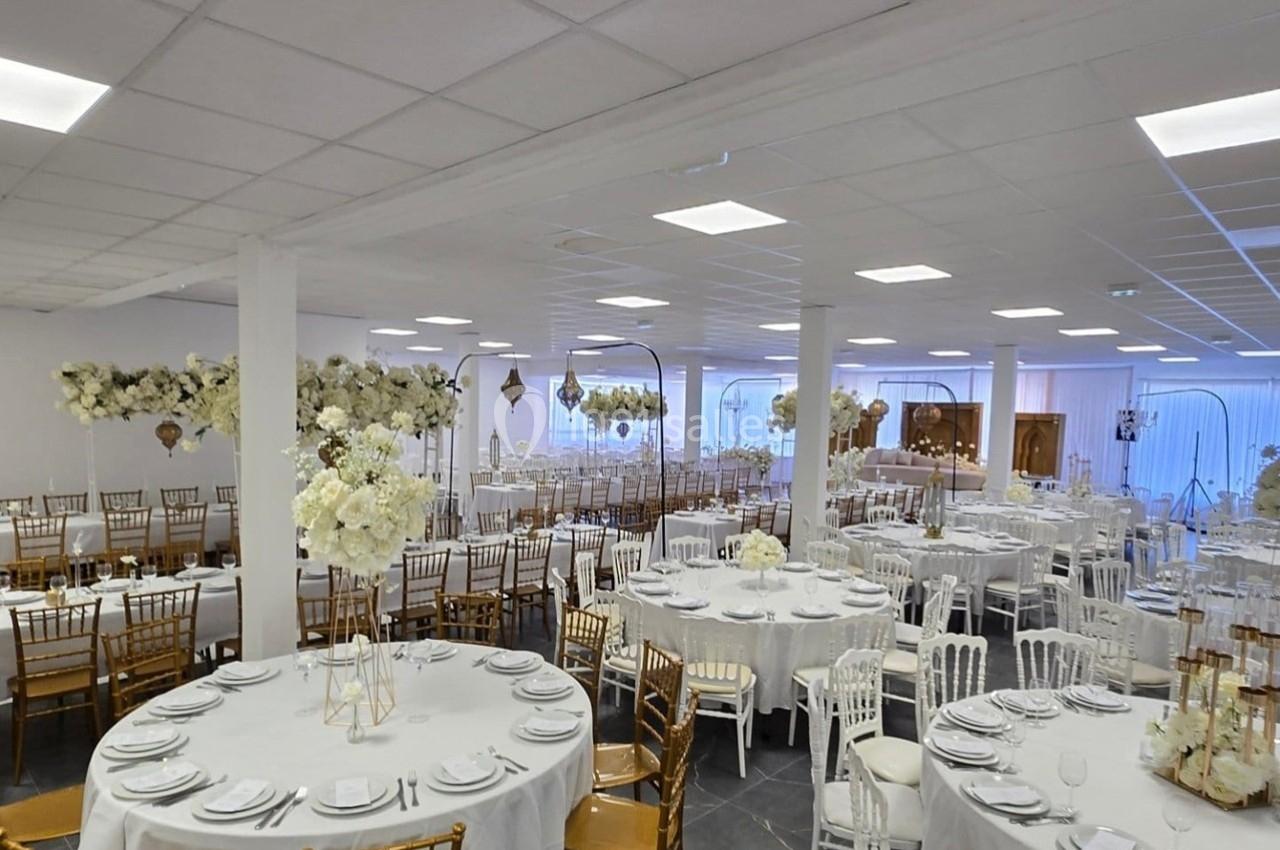 Salle de réception lumineuse avec tables rondes dressées, chaises blanches et dorées, et décorations florales blanches.