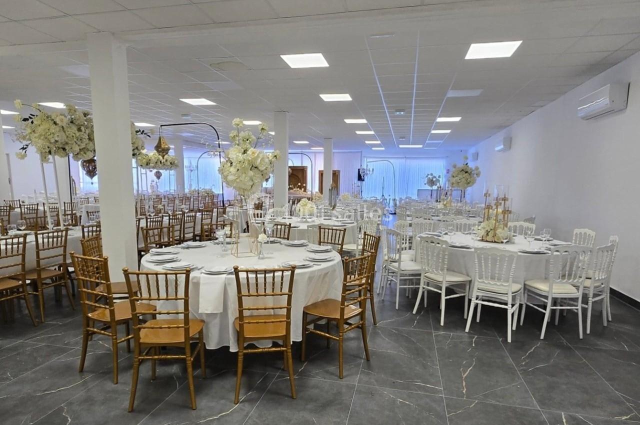 Salle de réception élégante avec tables rondes dressées, chaises dorées et blanches, et décorations florales blanches.