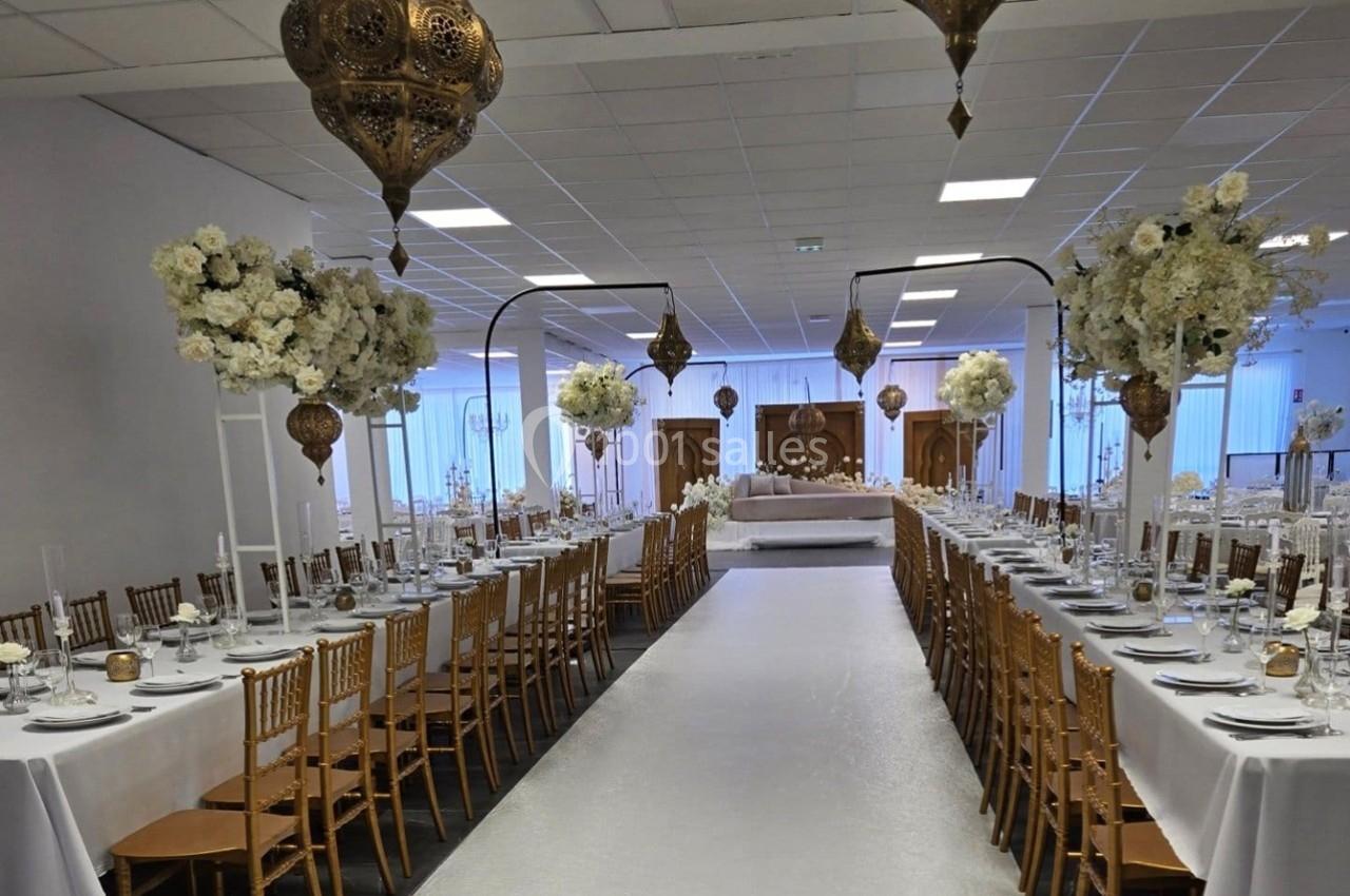 Salle de réception décorée avec des tables alignées, nappes blanches, chaises dorées, fleurs et lanternes suspendues.