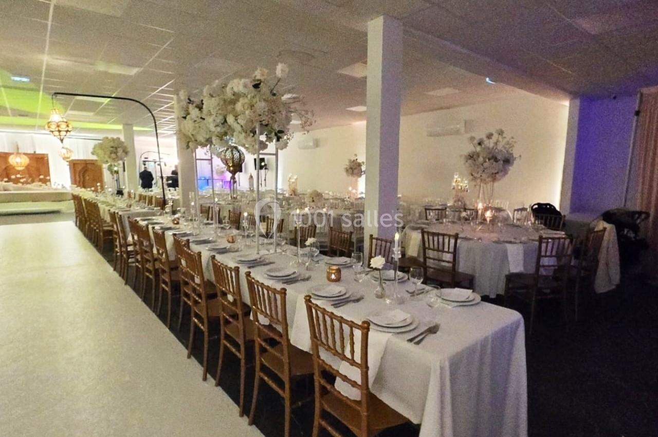 Grande salle de réception décorée avec des tables alignées, nappes blanches, chaises en bois et arrangements floraux.