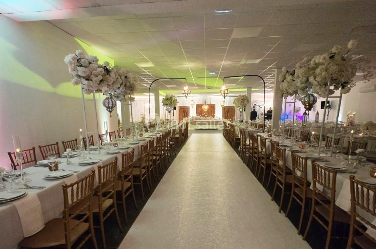 Salle de réception décorée avec des tables alignées, chaises dorées et arrangements floraux blancs sur un long tapis central.