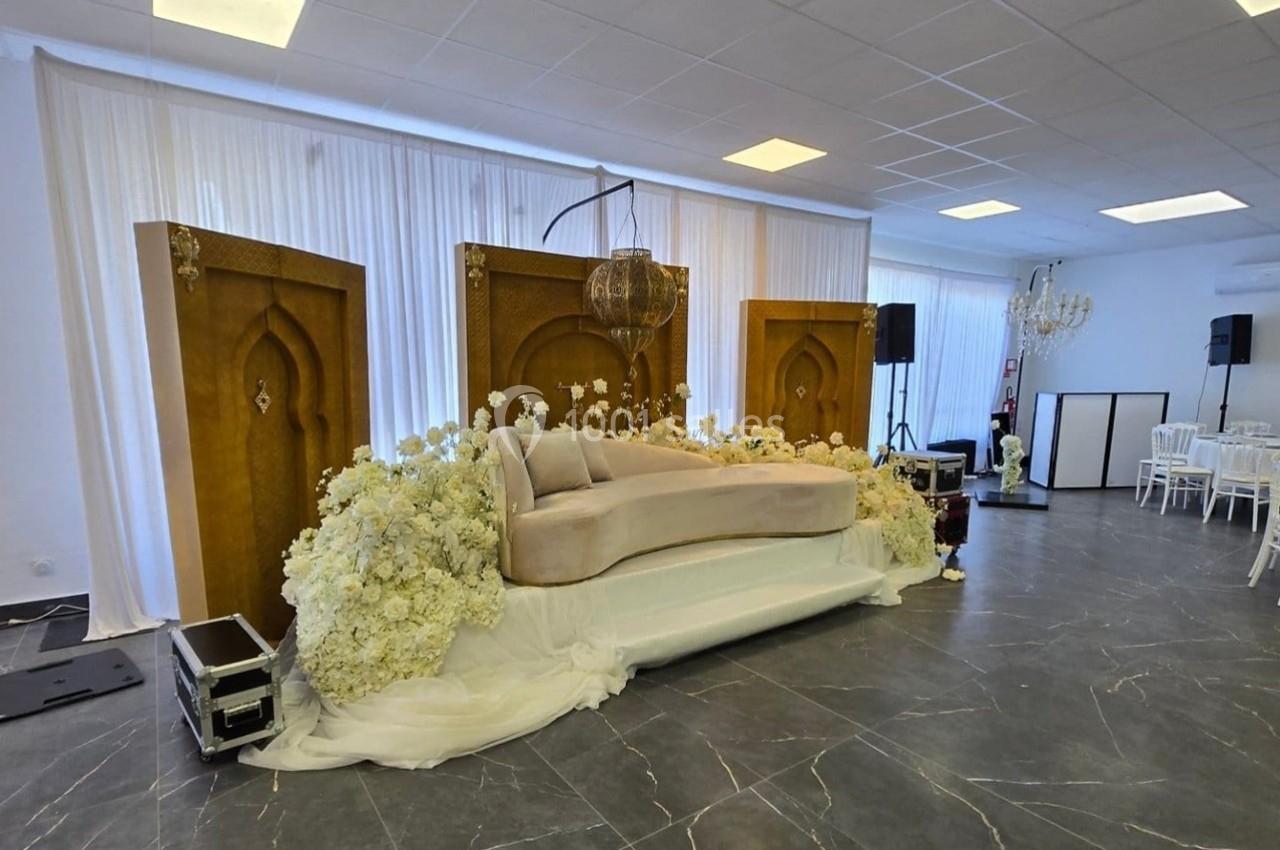 Canapé beige entouré de fleurs blanches, placé devant des panneaux dorés décoratifs dans une salle de réception.