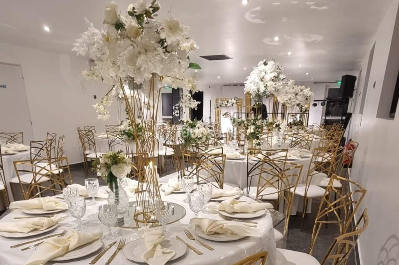 Salle de réception décorée avec des tables rondes, nappes blanches, chaises dorées et centres de table fleuris.
