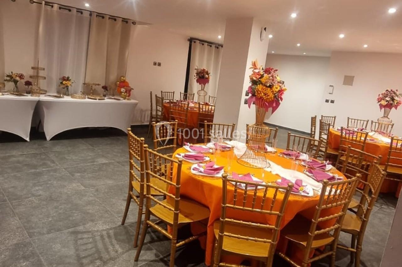 Salle décorée pour un événement avec tables rondes, nappes orange, chaises dorées et compositions florales colorées.