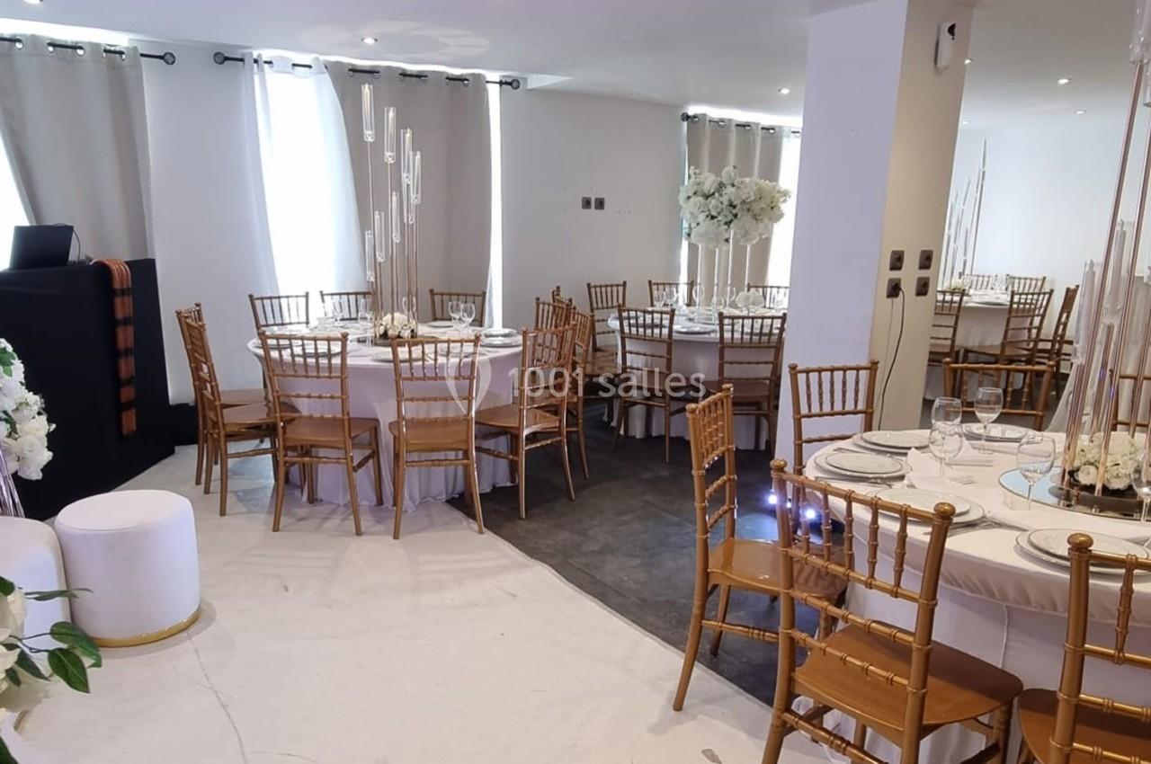 Salle décorée pour un événement avec des tables rondes, nappes blanches, chaises dorées et arrangements floraux blancs.