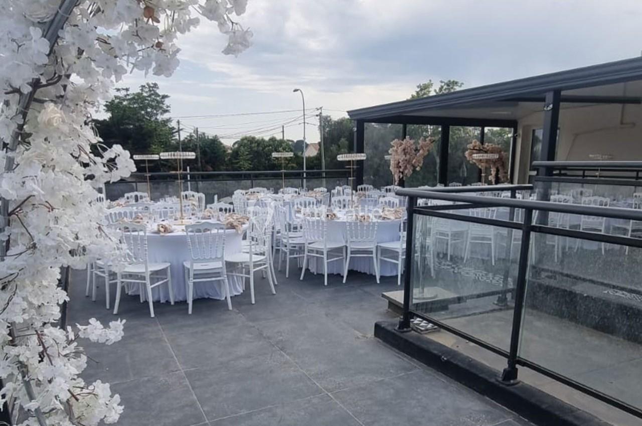 Salle extérieure aménagée pour un événement, avec des tables rondes dressées et des chaises blanches sous un ciel…