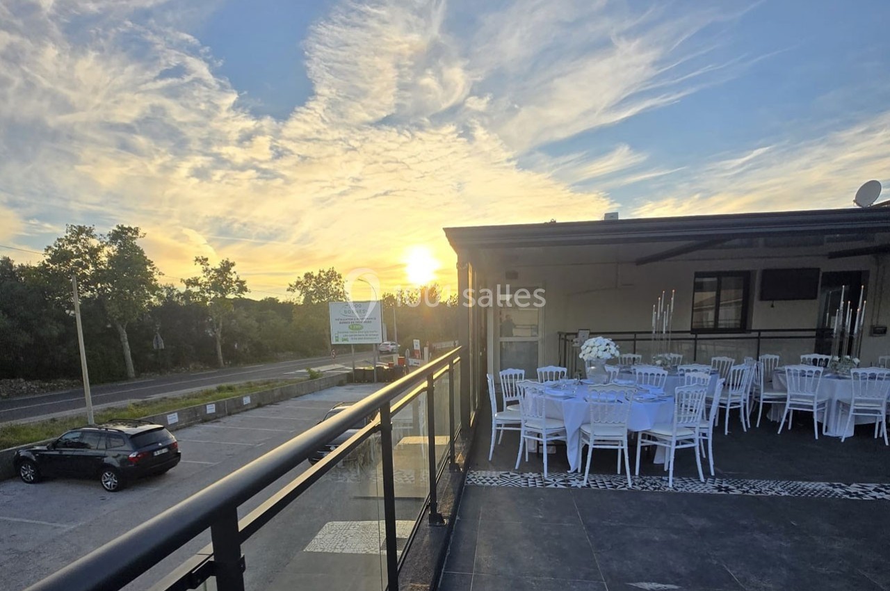 Terrasse aménagée avec des tables et chaises blanches, vue sur un parking et un coucher de soleil à l'horizon.