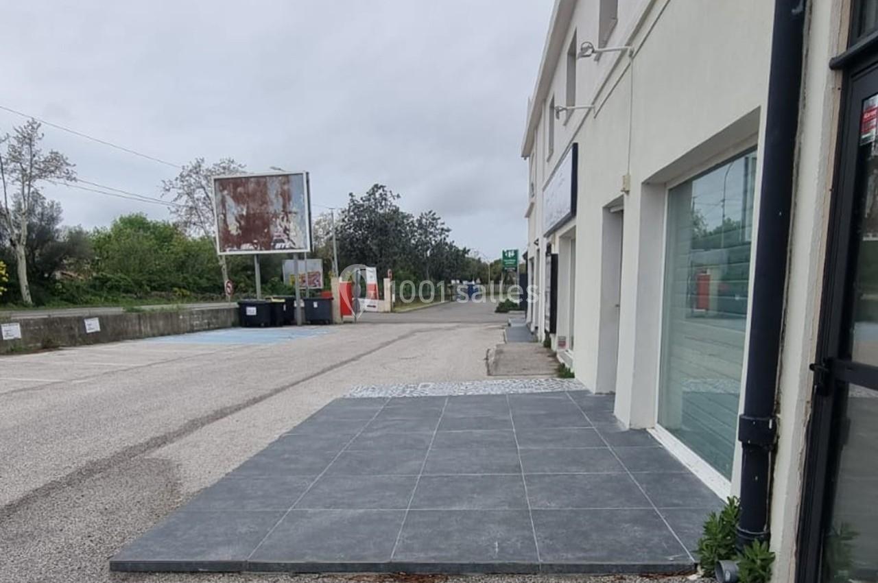 Trottoir en carrelage gris devant un bâtiment blanc, longeant une route bordée d'arbres et de panneaux.