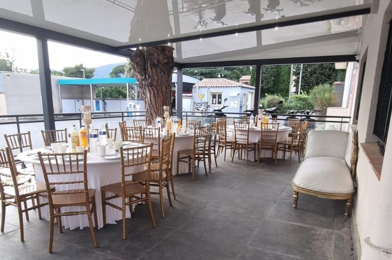 Salle extérieure couverte avec tables rondes dressées, chaises en bois et vue sur un espace extérieur arboré.