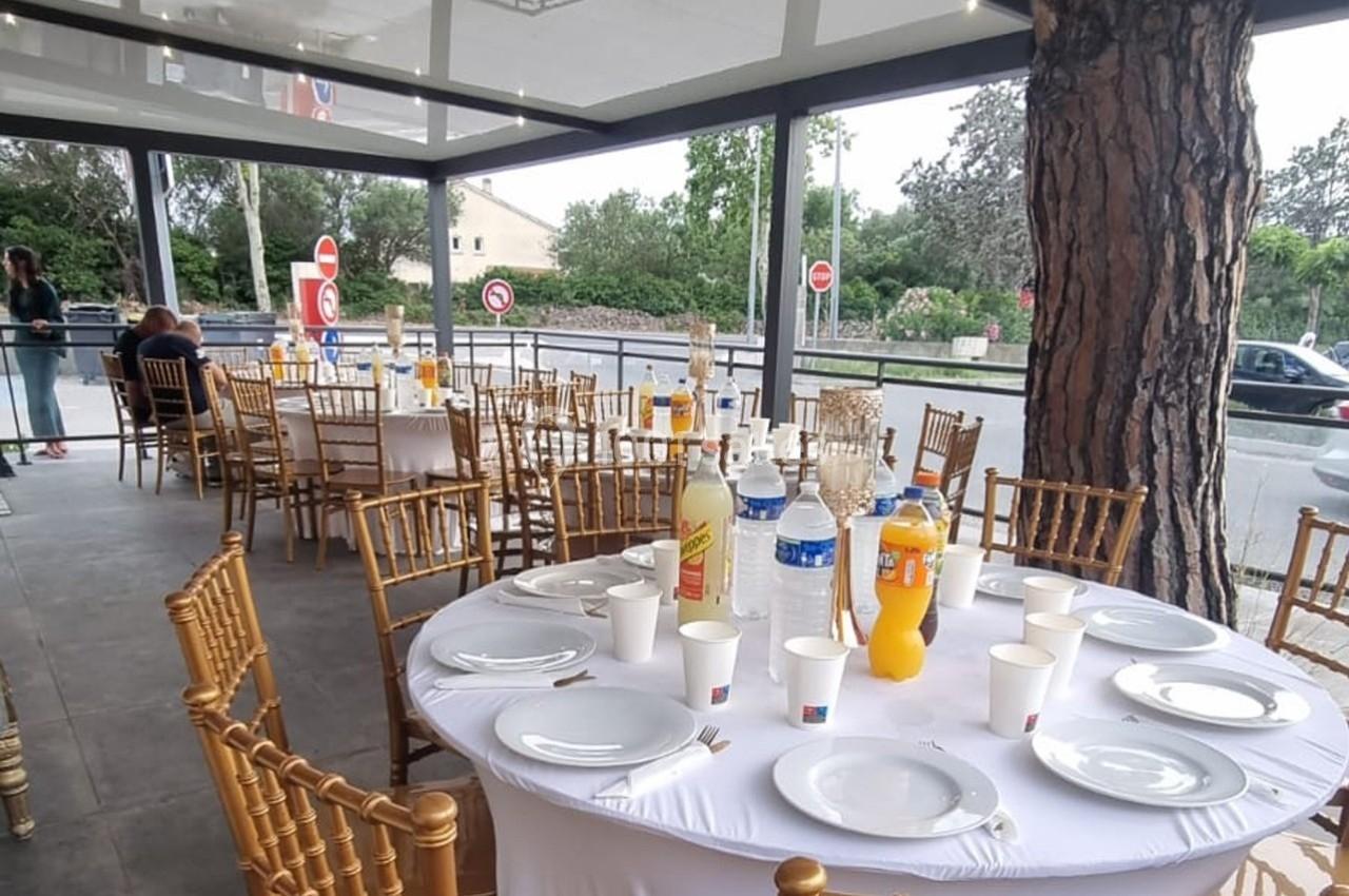 Tables rondes dressées avec nappes blanches, chaises dorées et boissons, dans un espace extérieur couvert.