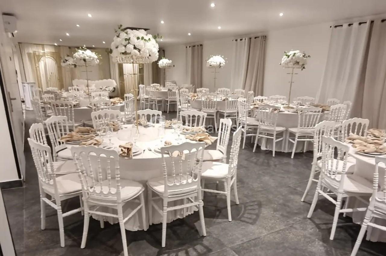 Salle de réception élégamment décorée avec des tables rondes, nappes blanches, chaises blanches et centres de table fleuris.