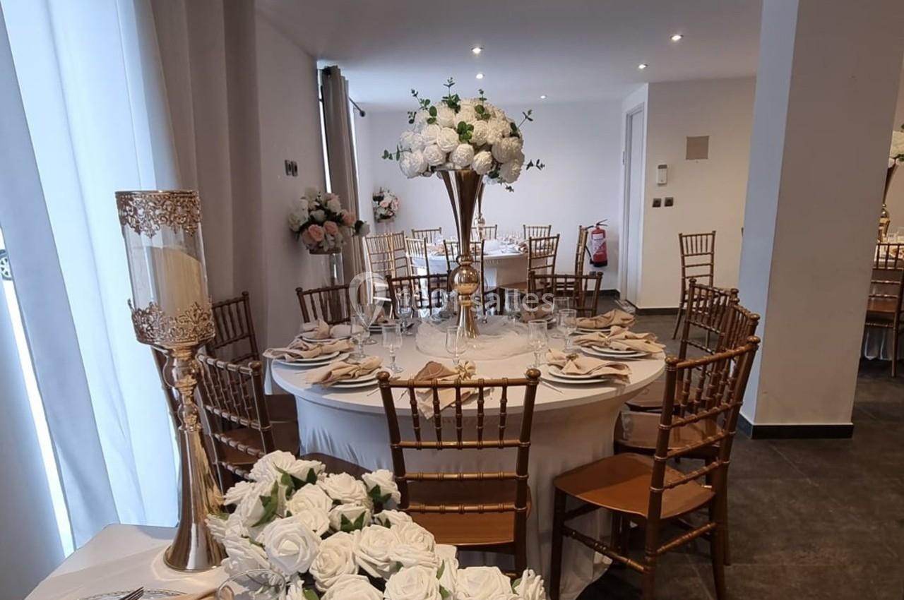 Salle décorée pour un événement avec tables rondes, chaises dorées, nappes blanches et centres de table fleuris.