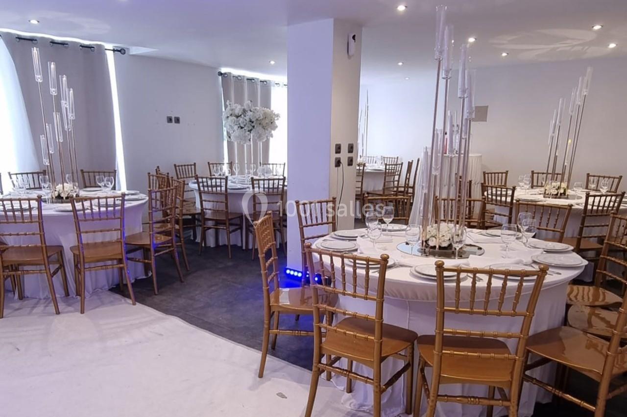 Salle de réception décorée avec des tables rondes, nappes blanches, chaises dorées et centres de table avec bougies.