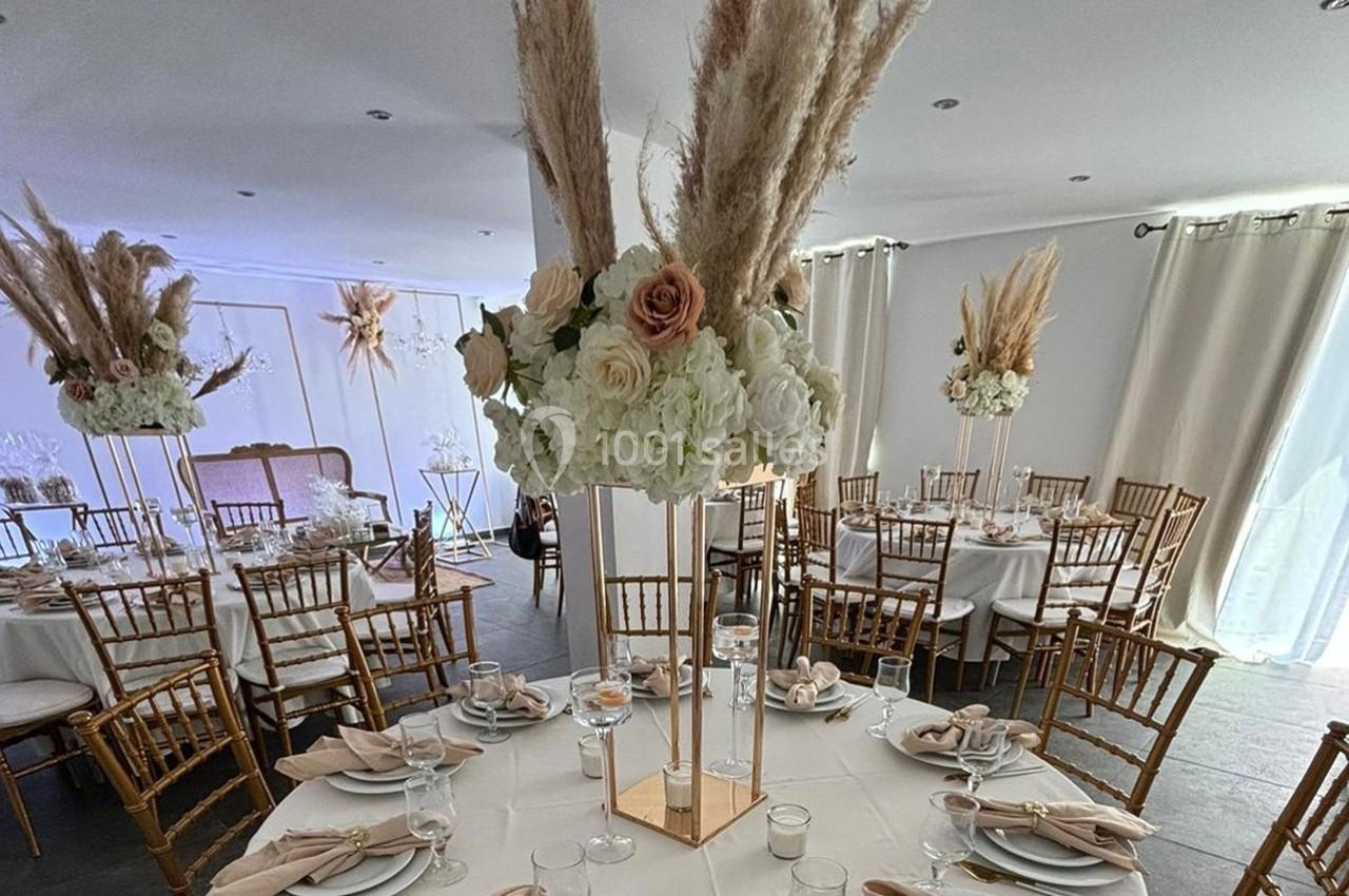 Salle de réception décorée avec des tables rondes, nappes blanches, chaises dorées et centres de table floraux élégants.