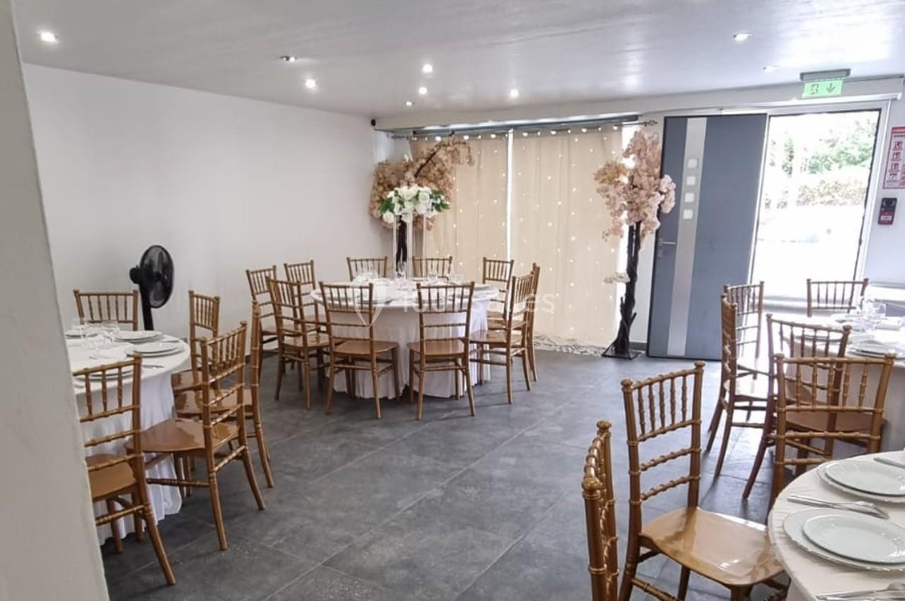 Salle de réception lumineuse avec tables rondes dressées, chaises en bois et décor floral près d'une baie vitrée.