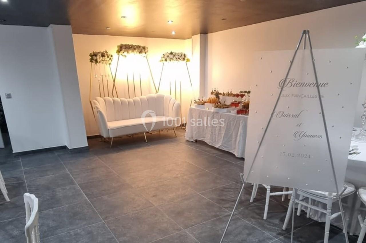 Salle décorée pour un événement avec canapé blanc, panneau de bienvenue et table de boissons sur fond lumineux.