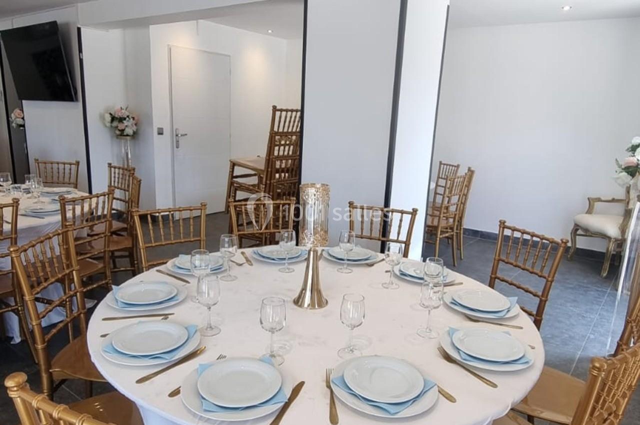 Salle décorée avec des tables rondes dressées, chaises dorées et centre de table avec bougie, ambiance élégante.