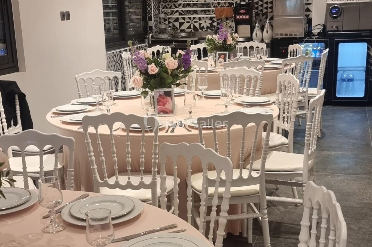Salle décorée pour un événement avec des tables rondes dressées, nappes roses, chaises blanches et bouquets de fleurs.