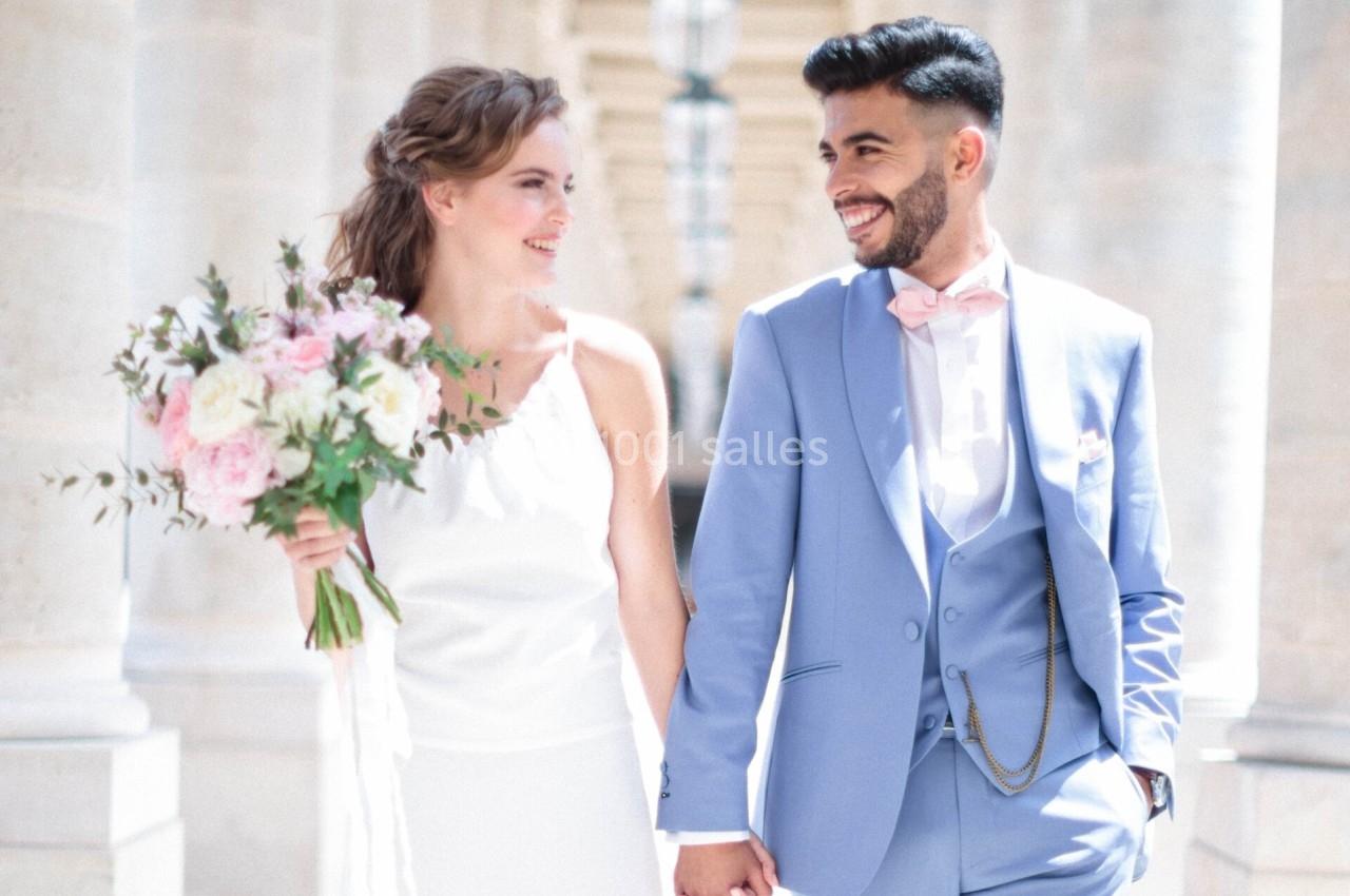 Un couple souriant en tenue de mariage marche main dans la main dans un lieu lumineux avec des colonnes en arrière-plan.