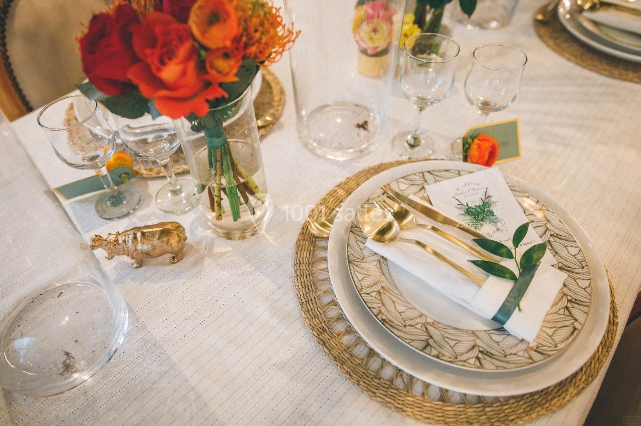 Table dressée avec des assiettes à motifs, couverts dorés, serviette nouée, fleurs rouges et orange en décoration.