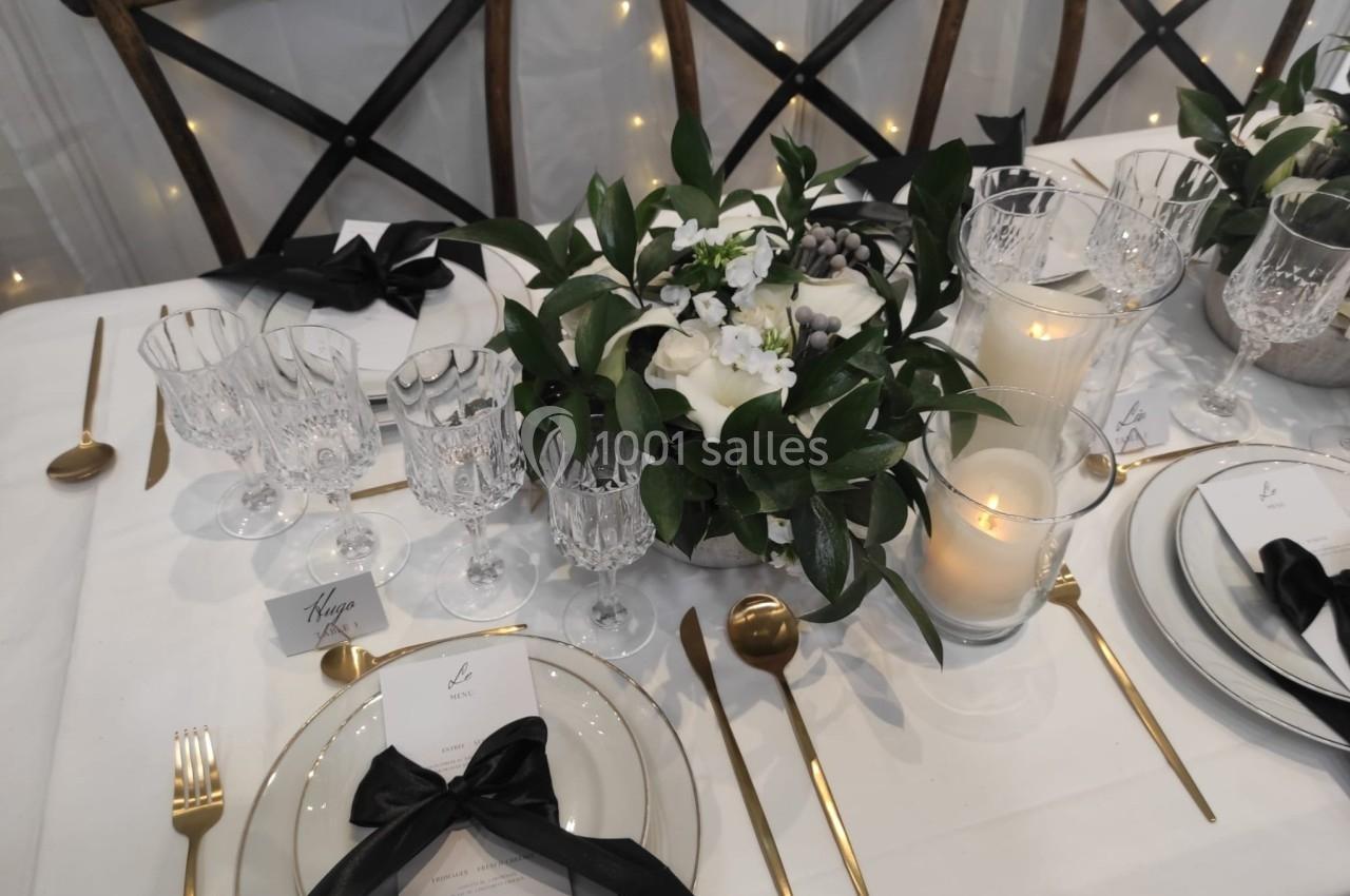 Table élégamment dressée avec vaisselle raffinée, couverts dorés, bougies et centre de table floral blanc et vert.