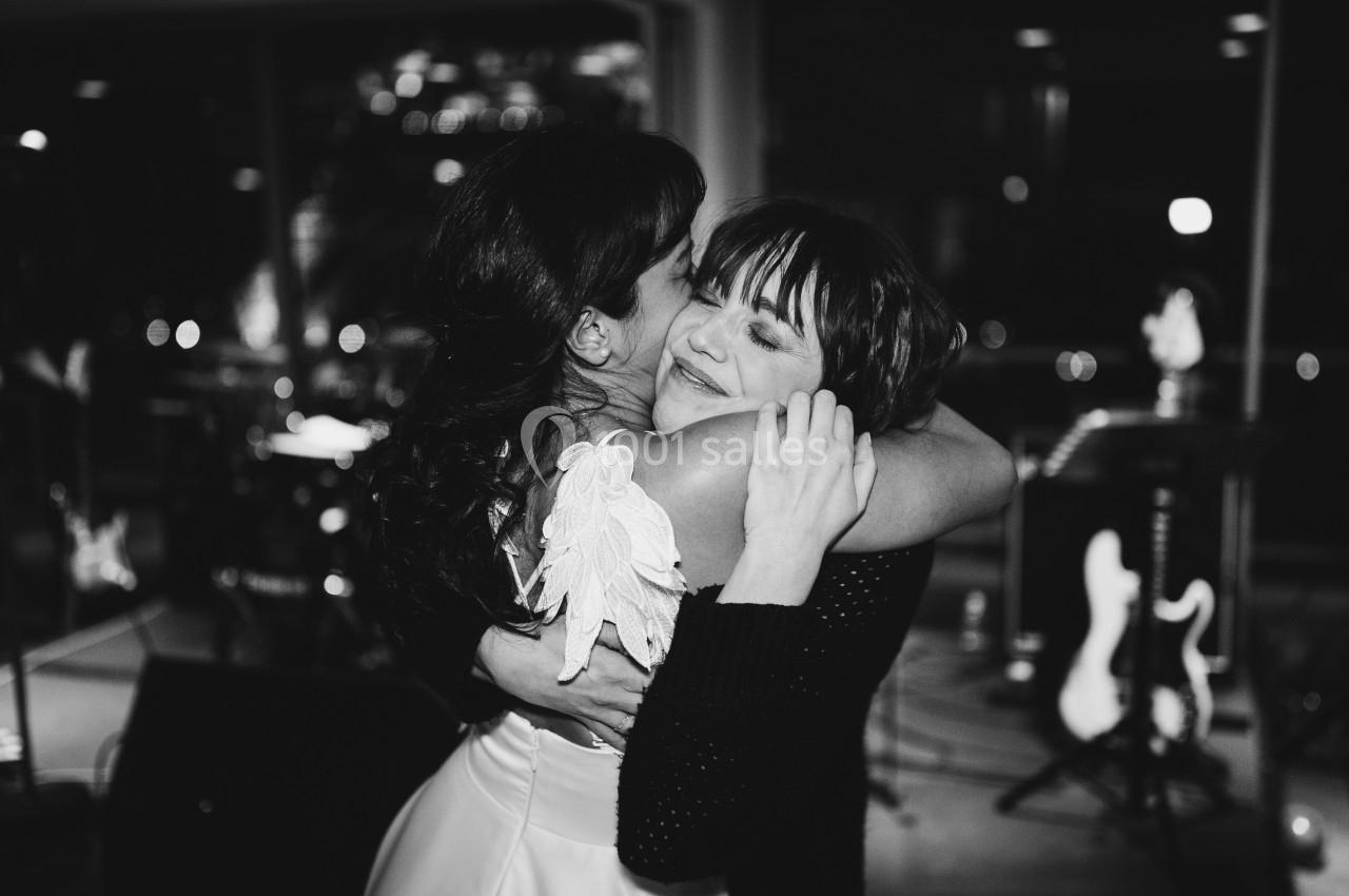 Deux femmes s'enlacent chaleureusement dans une ambiance festive, avec des instruments en arrière-plan.