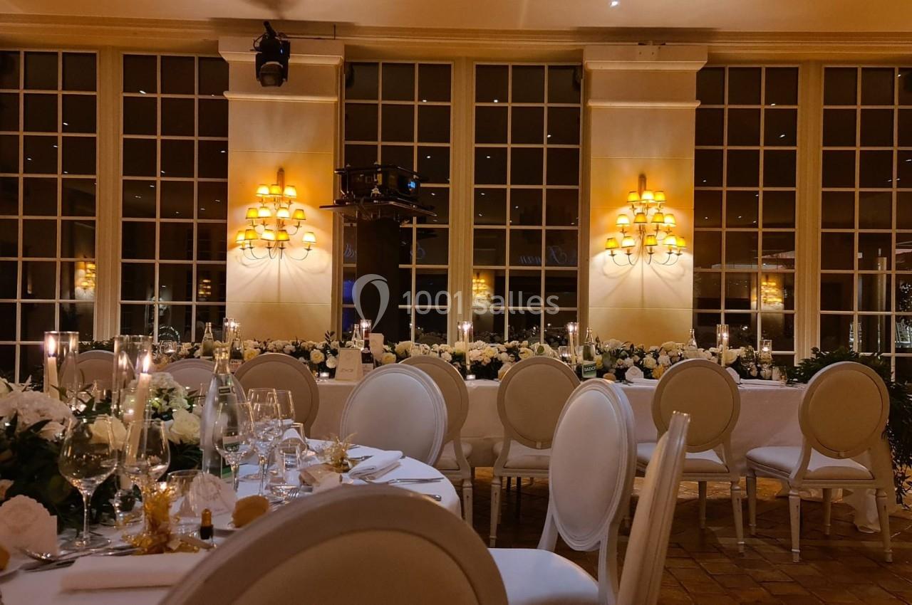 Salle de réception élégante avec tables dressées, chaises blanches et décorations florales sous un éclairage chaleureux.