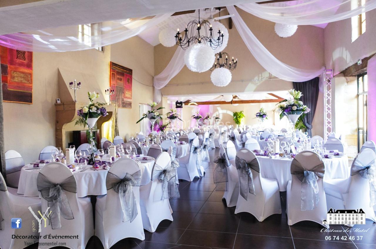 Salle de réception décorée avec des tables rondes, nappes blanches, chaises ornées de nœuds gris et éclairage tamisé.