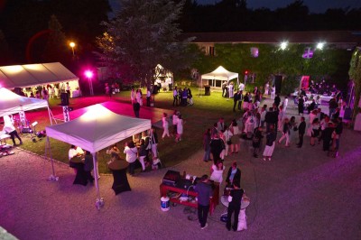 Allée gravillonnée bordée de lanternes blanches menant à un jardin arboré, vue depuis un portail en fer forgé.