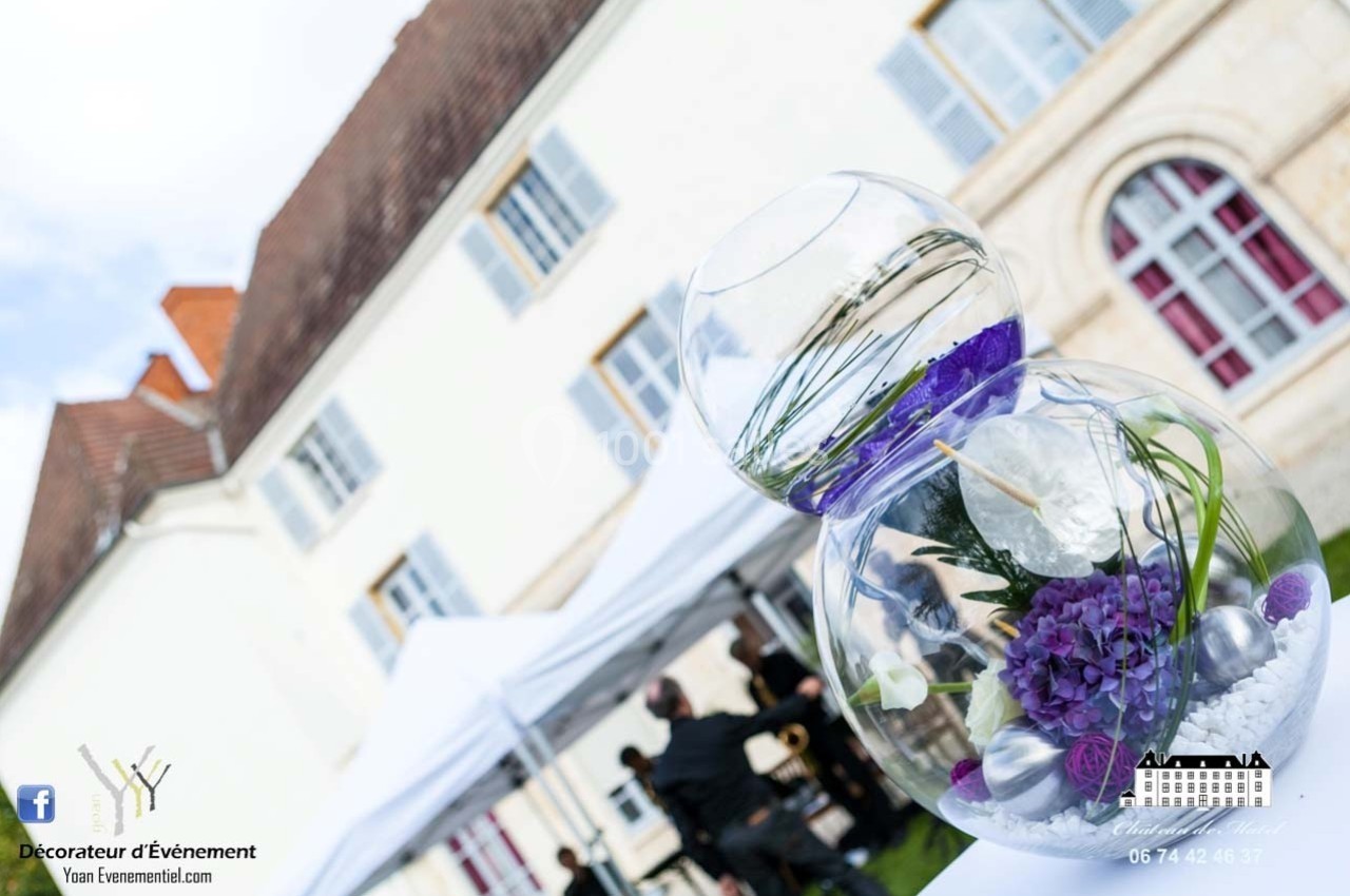 Composition florale violette dans un vase en verre devant une grande bâtisse avec des tentes blanches en arrière-plan.