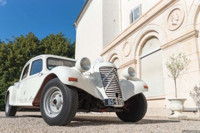 Allée gravillonnée bordée de lanternes blanches menant à un jardin arboré, vue depuis un portail en fer forgé.