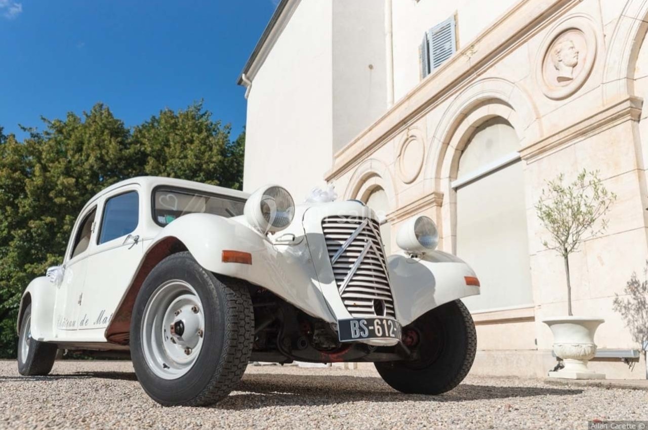 Voiture ancienne blanche stationnée devant un bâtiment clair avec des arches et des arbres en arrière-plan.