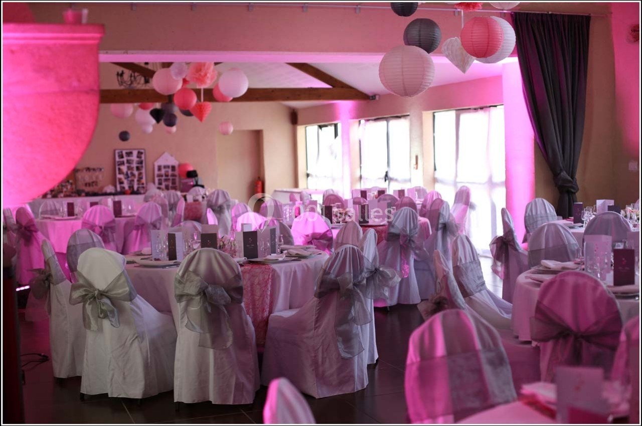 Salle de réception décorée pour un événement, avec des tables dressées, des chaises couvertes et des lanternes suspendues.
